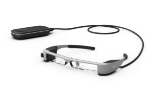 Imagen de las gafas de realidad aumentada usadas por el sistema Retiplus, las EPSON Moverio BT-300. En ella se observa estas gafas, con sus cristales totalmente transparentes, la microcámara incorporada en la patilla derecha, y el mando a distancia