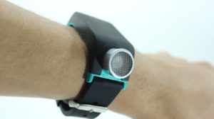 Imagen en la que se observa la pulsera Sunu Band en la muñeca de una persona. La pulsera es de color negro, a excepción de su parte inferior, que en este modelo es de color azul claro, y del sensor de ultrasonido que es plateado.
