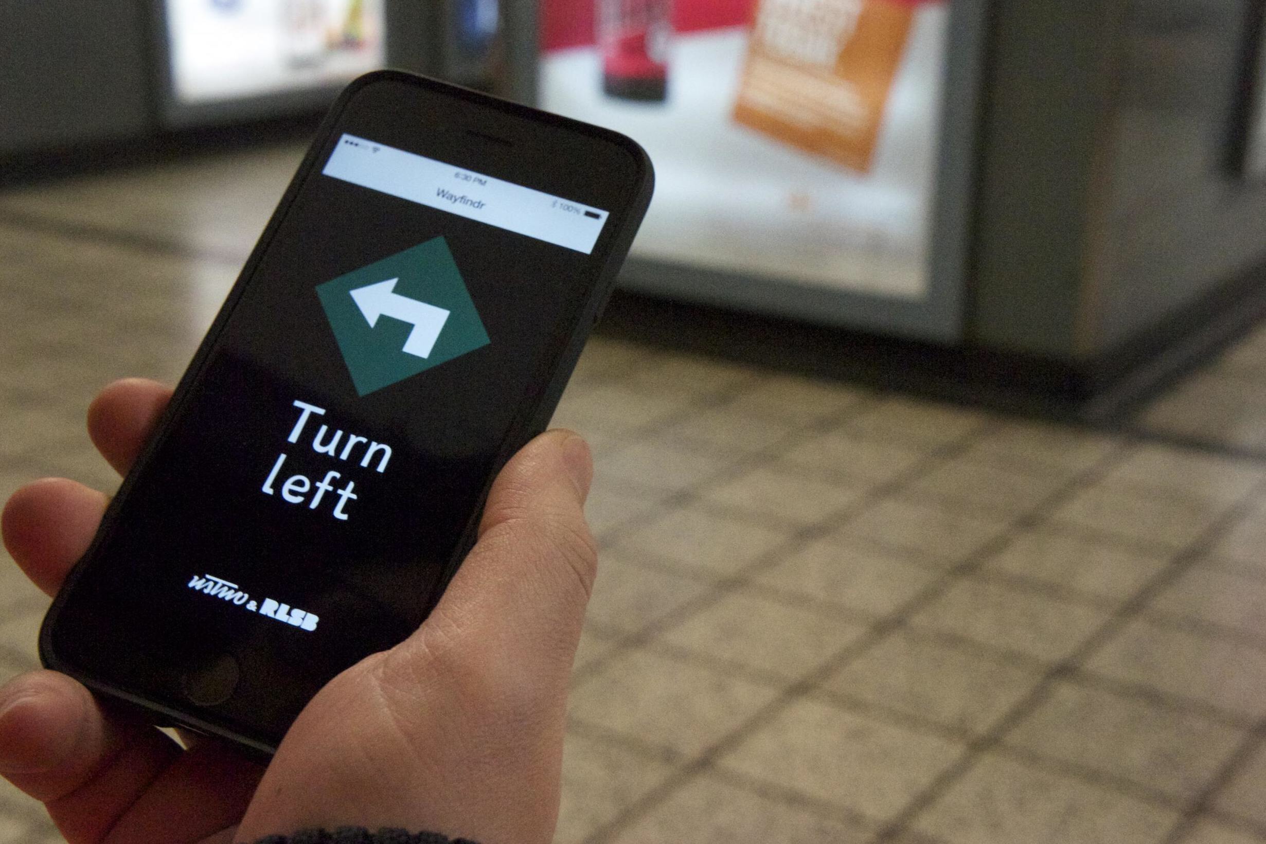 Imagen en la que aparece la mano de una persona empleando la aplicación Wayfindr en un smartphone. En la pantalla de éste aparece un mensaje con las palabras “Turn left” (gire a la izquierda) junto con una señal parecida a las de tráfico que indica que giremos en esa dirección. El fondo del móvil es negro, las letras son blancas y la señal es blanca sobre un fondo verde oscuro.