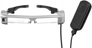 Imagen de las smartglasses de realidad aumentada empleadas en el sistema Retiplus, las EPSON Moverio BT-350. Las gafas, de aspecto futurista pero sin llegar a la extravagancia, son de color gris y negro, tienen unas lentes totalmente transparentes, una microcámara incorporada en la patilla derecha y sus patillas son graduables para poder adaptarse a los distintos tamaños de cabeza. En la imagen también aparece El controlador que permite interactuar con el sistema. Éste es de color negro, va unido por medio de un cable a las gafas, y en su interior se encuentran la batería y el procesador.