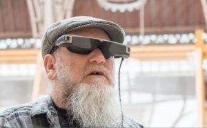 Imagen en la que se observa un hombre llevando las smartglasses para baja visión OxSight Prism. Las gafas son de color negro y gris metalizado, llevan la cámara integrada en la patilla derecha y engloban por completo a los ojos, ciñéndose por completo a la forma de la cara, sin dejar aberturas en los laterales. Los cristales son oscuros pero transparentes, cuentan en su interior con unas pantallas micro-OLED también transparentes, y de la patilla izquierda surge un cable que, aunque no se aprecia en la imagen, va a un controlador/procesador de color negro que incluye la unidad de procesamiento del sistema, la batería y los controles de las distintas funciones del equipo. Las gafas tienen una estética futurista pero sin llegar a ser muy aparatosas.