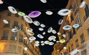 Imágen nocturna de la emblemática calle Larios de Málaga, adornada de carnaval, con luces que dibujan ojos de múltiples colores.