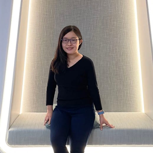 La imagen muestra a una mujer, con rasgos asiáticos La C_R_O y Doctora Florence Chan, posando para la fotografía. Ella está de pie con una postura relajada, ligeramente inclinada hacia un lado, con su mano derecha descansando sobre su cadera y la izquierda colgando junto a su cuerpo. Lleva gafas con montura y su cabello es largo, liso y de color oscuro, cayendo libremente sobre sus hombros. Viste una blusa de manga larga de color negro y pantalones ajustados también negros. Lleva lo que parece ser una pulsera o reloj azul en su muñeca izquierda. La mujer se encuentra en un espacio interior con una iluminación suave y uniforme. Detrás de ella, hay una estructura que parece ser una silla alta con un respaldo grande y rectangular, que está iluminado por detrás, creando un efecto de halo alrededor de la estructura.