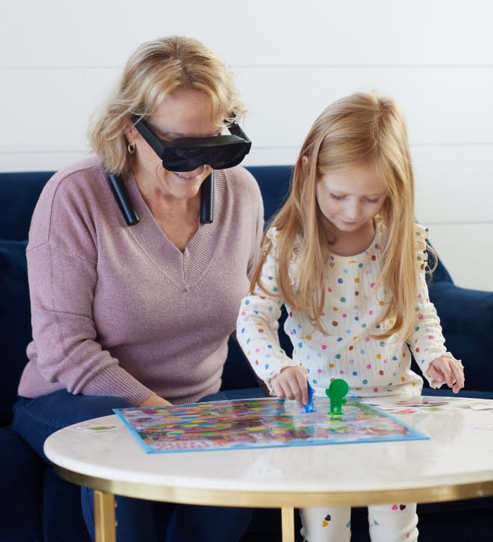 En la imagen hay una mujer mayor y una niña pequeña sentadas en una mesa redonda jugando un juego de mesa. La mujer lleva unas gafas Esight y parece estar disfrutando del juego con la niña. Ambas están sonriendo y concentradas en el juego. La habitación tiene una decoración moderna y minimalista.