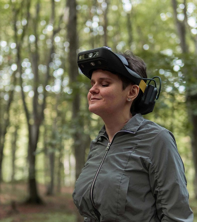 La imagen muestra a una mujer en un bosque. Ella lleva un dispositivo en la cabeza que es un visor Lumen. También tiene puestos unos auriculares protectores. La mujer tiene los ojos cerrados y una expresión serena en su rostro. El fondo está desenfocado, pero se pueden ver árboles y la luz que se filtra a través del follaje.