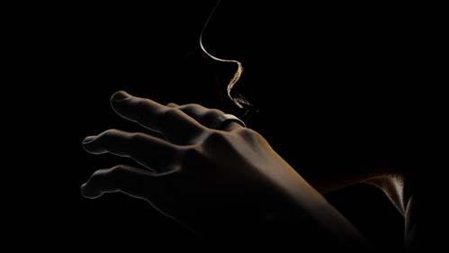 La imagen muestra una mano humana vista de perfil contra un fondo oscuro. La mano está elevada con la palma hacia abajo y los dedos ligeramente curvados, como si estuviera en reposo o tal vez en el proceso de realizar un gesto. Lo más destacado de la imagen es una delgada línea de luz que parece emanar o interactuar con el dedo índice de la mano, sugiriendo algún tipo de tecnología o efecto especial. La luz tiene una forma ondulante y sinuosa que recuerda a una corriente suave de humo o vapor, y se destaca contra el fondo oscuro, lo que da un aspecto etéreo y delicado a la composición. La fotografía parece ser muy estilizada, con un enfoque en la línea de luz, lo que podría indicar que se trata de una imagen conceptual relacionada con tecnología wearable como podría ser un anillo inteligente.