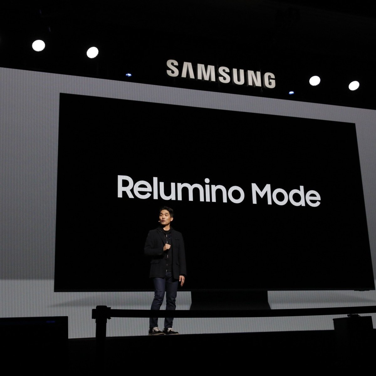 La imagen muestra a una persona de pie en un escenario frente a una gran pantalla con la palabra "SAMSUNG" en la parte superior y "Relumino Mode" en el centro en un gran texto blanco. La persona parece estar dando una presentación o un discurso. El fondo de la pantalla es completamente negro, lo que hace que las letras blancas resalten significativamente. La persona está vestida con ropa casual formal: una chaqueta oscura sobre una camisa oscura y jeans, complementada con zapatos oscuros. La iluminación del escenario se centra en el presentador, con luces adicionales apuntando hacia la pantalla desde arriba. El tamaño y el diseño de la presentación sugieren que se trata de un evento importante, probablemente una conferencia o lanzamiento de producto de Samsung.