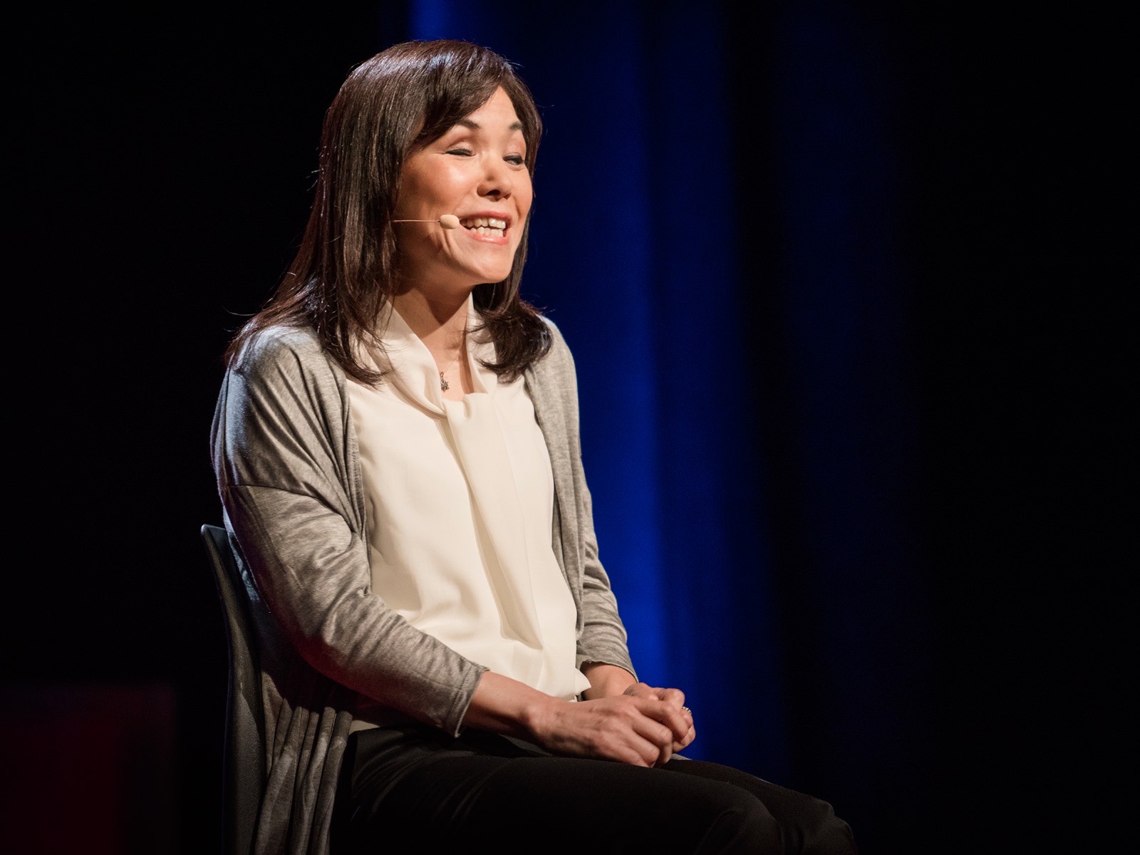 La imagen muestra a la Doctora Asakawa sentada en un escenario dando una charla TED. Está usando un micrófono de diadema. Lleva una chaqueta gris sobre una blusa de color claro, y pantalones oscuros. Su cabello es oscuro y llega hasta un poco más abajo de los hombros. La mujer parece estar sonriendo o hablando con expresión animada. El fondo es oscuro, probablemente para no distraer la atención del público y concentrarla en la oradora.