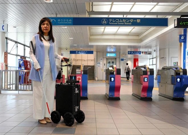 La imagen muestra a la Doctora Asakawa de pie, como esperando, frente a unos torniquetes de acceso en una estación de tren o metro. Ella tiene el cabello largo y lleva un conjunto de vestimenta clara con pantalones y un chaleco largo sin mangas. A su lado, hay una maleta AI Suitcase negra y robusta con varias ruedas motorizadas de aspecto robótico. La estación está marcada con señales en japonés y se ve el letrero de "Telecom Center Station". El área es espaciosa y bien iluminada, con un ambiente ordenado y limpio. En el fondo, se puede ver a una persona caminando y la entrada o salida de la estación de tren o metro.