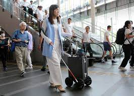 La imagen muestra a la Doctora Asakawa caminando por una estación de tren o metro, llevando consigo un bastón blanco. Está utilizando también una maleta guía con ruedas de aspecto tecnológicamente avanzado, AI Suitcase. La escena tiene lugar en un espacio interior amplio y bien iluminado, una terminal pública, con piso de baldosas y barandas de metal. Varias otras personas se pueden ver en el fondo, algunas subiendo escaleras mecánicas y otras caminando en distintas direcciones. La atmósfera es la de un lugar transitado, y todo el mundo parece estar ocupado con sus prisas diarias.