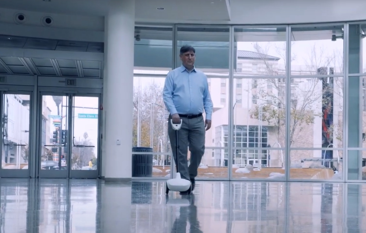 La imagen muestra a un hombre en un interior espacioso y luminoso, como podría ser un vestíbulo o una entrada pública. Está utilizando un dispositivo de movilidad Glide. La estructura de Glide consiste en un mango ergonómico unido a una estructura vertical que termina en una base con ruedas. El diseño sugiere funcionalidades avanzadas, con inteligencia artificial y sensores para ayudar en la navegación y la detección de obstáculos. La postura del hombre indica que él está caminando de forma autónoma y confiada, guiado por el dispositivo.