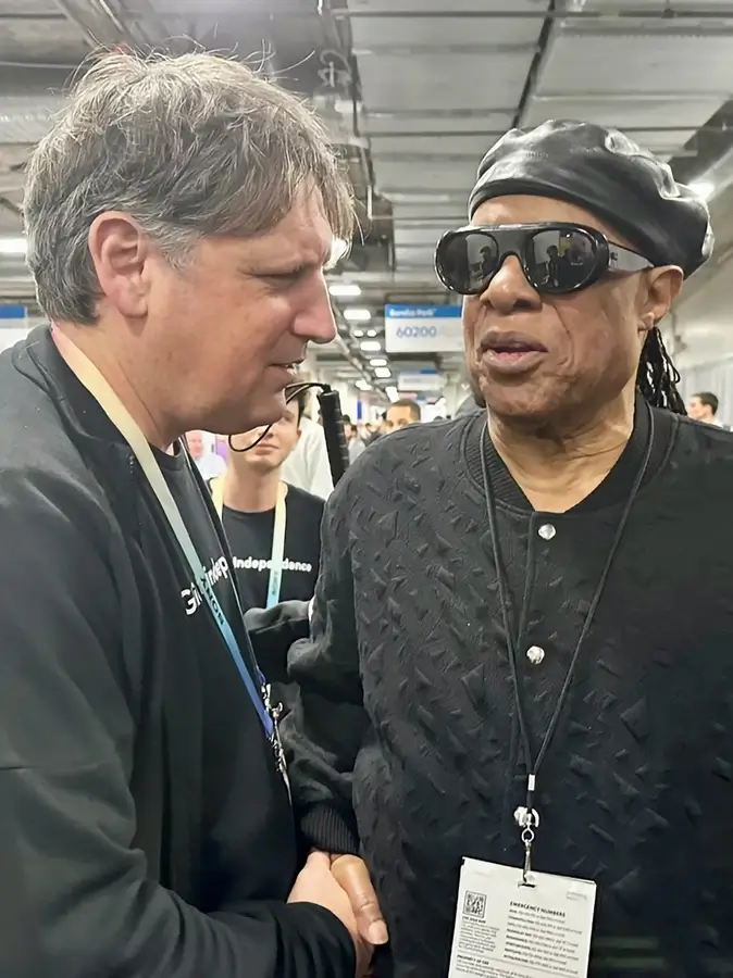 La imagen muestra a amos-miller-y stevie-wonder interactuando en el entorno de la conferencia tecnológica CES 2024. Amos, que es el hombre que está a la izquierda lleva una etiqueta de identificación que indica que está relacionado con la empresa Glidance y porta una camiseta de color claro con la frase "Glide into independence". El hombre de la derecha, que lleva gafas de sol oscuras y una boina de cuero, es Stevie Wonder. Ambos están estrechándose las manos en un gesto amistoso, posiblemente en el contexto de una demostración o discusión sobre el producto o tecnología mencionada anteriormente. La interacción entre ellos parece amena y cordial, con Stevie Wonder sonriendo ligeramente hacia el hombre de la izquierda, indicando una conversación positiva o un encuentro agradable.