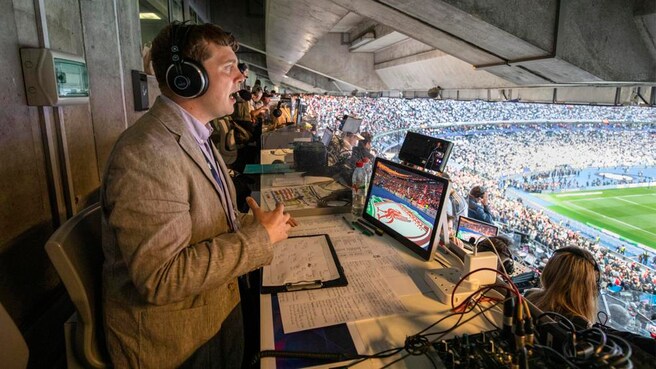 La imagen muestra una cabina de comentaristas deportivos en un estadio lleno de espectadores. Un comentarista está hablando al micrófono mientras lleva auriculares y se concentra en su tarea. La cabina está equipada con varias pantallas de computadora, documentos y equipos de audio. A través de la ventana, se puede ver claramente el campo de fútbol y una gran multitud de espectadores en las gradas. La atmósfera es animada, lo que sugiere que se está llevando a cabo un evento deportivo importante, posiblemente un partido de la UEFA Champions League o un evento similar. 