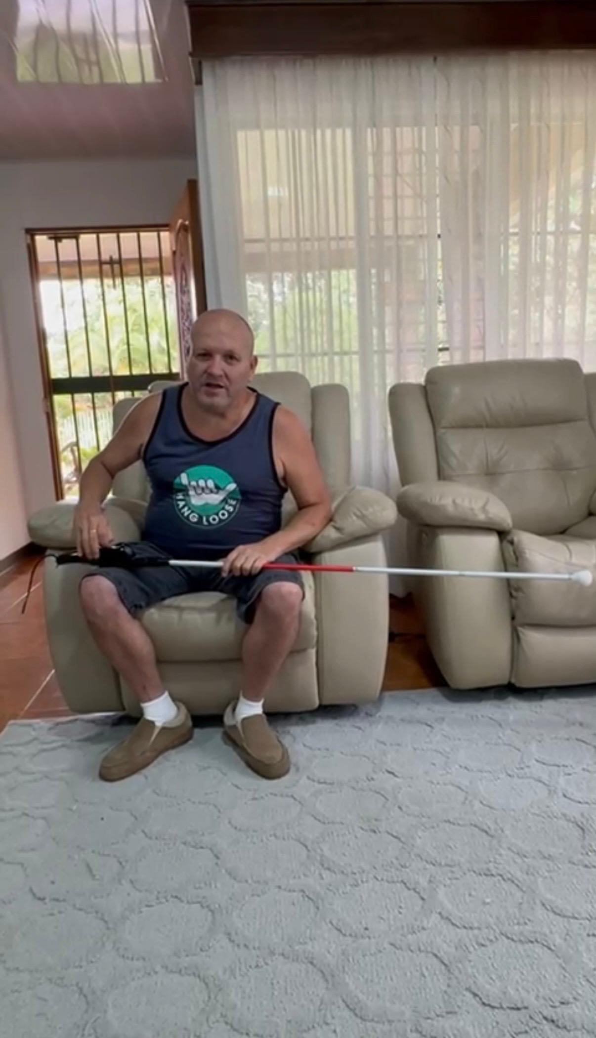 La imagen muestra a Víctor Hugo sentado en un sillón reclinable individual de color beige claro. Viste una camiseta sin mangas azul oscuro con un logo circular verde que muestra una mano haciendo el signo "shaka" y el texto "HANG LOOSE" debajo. Lleva pantalones cortos de color gris oscuro, calcetines blancos cortos y zapatos sin cordones de color marrón claro. Sostiene un bastón blanco con una sección roja y un mango negro. Su mano izquierda está en el mango negro del bastón. Mira directamente al frente. Detrás de él, a su izquierda, hay una puerta de madera con una reja que permite ver vegetación verde en el exterior. A su derecha, hay una ventana grande con cortinas blancas translúcidas, detrás de las cuales se ve más vegetación. Junto a Víctora su derecha, hay otro sillón reclinable individual idéntico y vacío. El suelo debajo de los sillones es de baldosas marrones. En primer plano, una alfombra gris claro con un patrón sutil de formas ovaladas o de nubes interconectadas cubre el suelo.