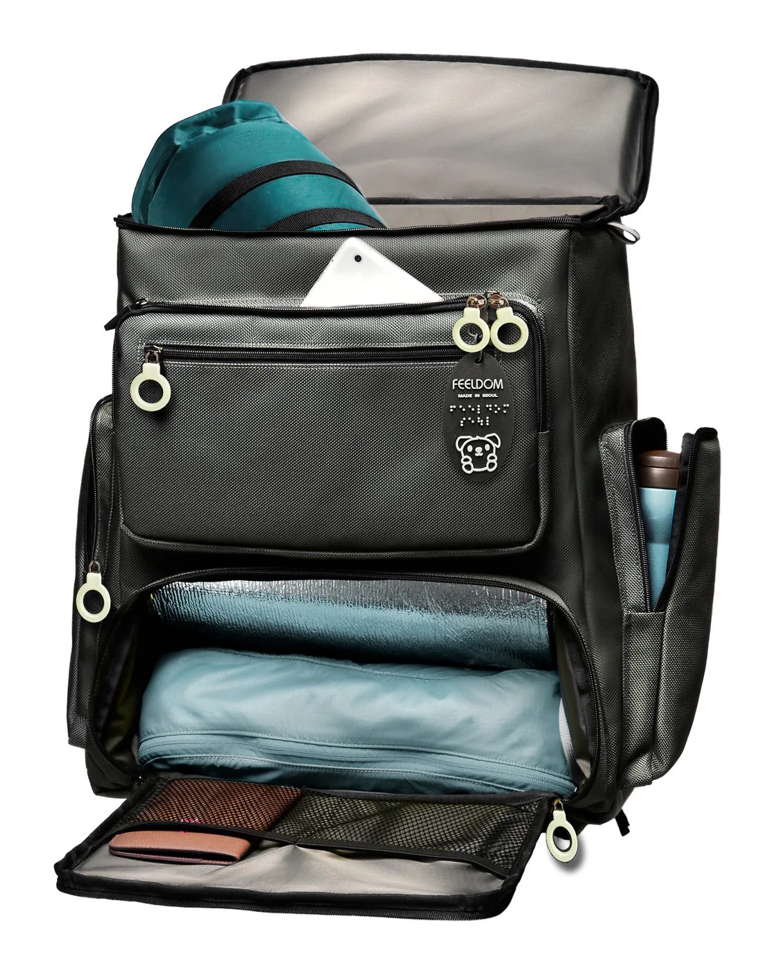 Vista frontal completa de la mochila cerrada. Presenta tres amplios compartimentos principales, dos de ellos con cremalleras dobles en posición central y uno inferior más ancho. A los lados se aprecian bolsillos verticales con cierre de cremallera. En la base frontal figura el logotipo bordado de FEELDOM sobre una etiqueta negra. El color predominante es gris grafito con herrajes claros de gran visibilidad. La estructura es rígida y mantiene su forma, incluso vacía, lo que evidencia su diseño autoestable.