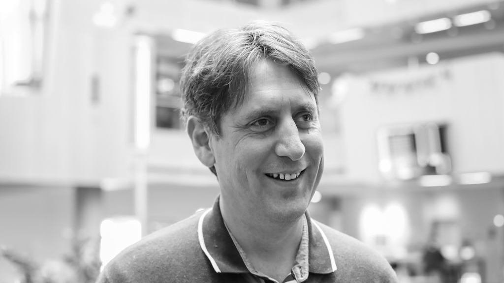 La imagen muestra a Amos Miller en un retrato en blanco y negro, en un entorno interior moderno y luminoso. Aparece ligeramente de perfil, con una expresión relajada y una sonrisa natural, vestido con una camiseta tipo polo. El fondo desenfocado sugiere un espacio profesional o académico, y la imagen transmite cercanía, serenidad y un perfil claramente vinculado al ámbito de la investigación y la innovación.