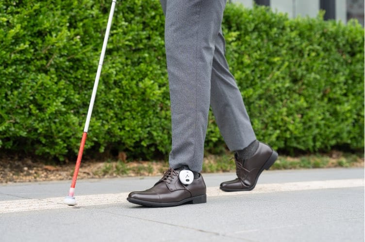La imagen muestra a una persona caminando por una acera urbana, captada desde la cintura hacia abajo, en un entorno exterior limpio y ordenado. Viste pantalón de vestir gris y zapatos clásicos de piel marrón, lo que refuerza la idea de un uso cotidiano y no deportivo del sistema. En la mano izquierda se aprecia claramente un bastón blanco, utilizado de forma activa para explorar el terreno delante de los pies. En el lateral del zapato derecho, a la altura del tobillo, se distingue el dispositivo Ashirase: un módulo compacto, circular y de color claro, firmemente acoplado al calzado mediante un soporte discreto. Su tamaño es reducido y su integración visual resulta poco intrusiva, permitiendo que el zapato conserve su estética convencional.
