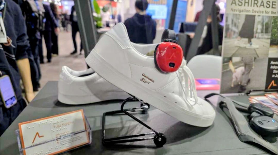 La imagen muestra un zapato deportivo blanco, de estilo urbano y diseño limpio, expuesto sobre un soporte metálico en un entorno de feria o exposición tecnológica. En el lateral exterior del calzado, a la altura del tobillo, se aprecia claramente el dispositivo Ashirase: un módulo compacto, de forma redondeada, con carcasa roja brillante y detalles en negro, firmemente acoplado al zapato. El dispositivo destaca visualmente por contraste cromático frente al blanco del calzado y parece diseñado para pasar desapercibido una vez integrado en el uso cotidiano. Su posición sugiere que transmite información directamente al pie mediante vibraciones, sin interferir con el movimiento natural al caminar. En el fondo de la imagen se distinguen otros elementos propios de un stand tecnológico: paneles informativos con el nombre “ASHIRASE Navigation Device”, material promocional y personas desenfocadas transitando por el recinto, lo que refuerza la idea de que se trata de una demostración pública del producto, probablemente en una feria internacional de tecnología.