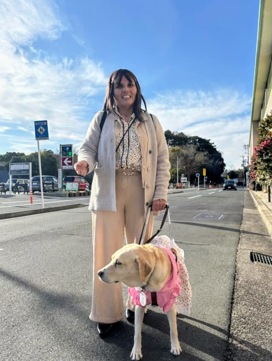 La imagen muestra a Wendi de pie en una vía urbana amplia y despejada, en un entorno exterior luminoso, con cielo azul y nubes altas. La escena está tomada a plena luz del día y transmite una sensación de calma y normalidad cotidiana. Wendi viste ropa clara y cómoda, con un conjunto en tonos beige y crema que refuerza una imagen natural y cercana. Lleva el cabello suelto y una expresión relajada, ligeramente sonriente, orientada hacia el frente. A su lado camina su perra guía, Paola, una labradora de color claro que avanza con paso firme y seguro. Paola viste un arnés y una prenda rosa que la hace claramente identificable y visible, y mantiene una postura atenta y concentrada, propia de un perro guía en servicio. La correa y el arnés evidencian el vínculo funcional y de confianza entre ambas, así como el trabajo coordinado que realizan durante el desplazamiento. El entorno urbano incluye una calzada asfaltada, señalización vial y edificios a ambos lados, sugiriendo un trayecto habitual dentro de la ciudad.