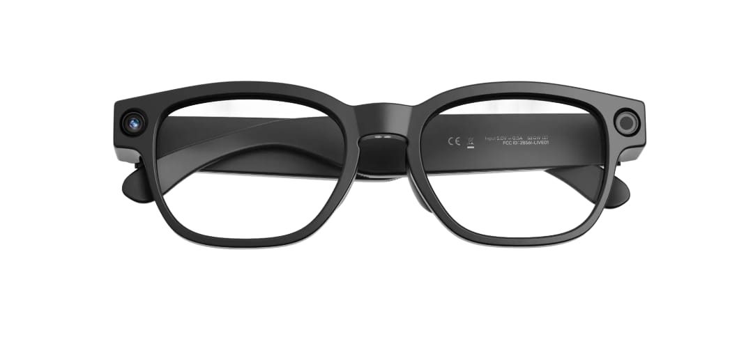 La imagen muestra unas gafas inteligentes negras de estilo clásico tipo wayfarer moderno, vistas en perspectiva lateral y ligeramente desde arriba, sobre un fondo completamente blanco. La montura tiene una forma rectangular con bordes suavemente redondeados, muy similar a unas gafas graduadas tradicionales, con un diseño sobrio y atemporal pensado para no llamar la atención. En la patilla izquierda, cerca de la bisagra, se aprecia claramente una cámara integrada, visible como un pequeño círculo con lente. La patilla es algo más gruesa de lo habitual, lo que indica que en su interior alberga componentes electrónicos, aunque sin romper la estética clásica. En la parte exterior de la patilla se distingue un logotipo pequeño y discreto. Los cristales son transparentes y la montura presenta un acabado negro mate, reforzando un aspecto elegante, funcional y cotidiano. Las patillas están abiertas, mostrando su curvatura natural hacia el extremo que apoya en la oreja. En conjunto, el diseño es discreto y equilibrado, pensado para llevarse durante todo el día y pasar por unas gafas normales a simple vista.