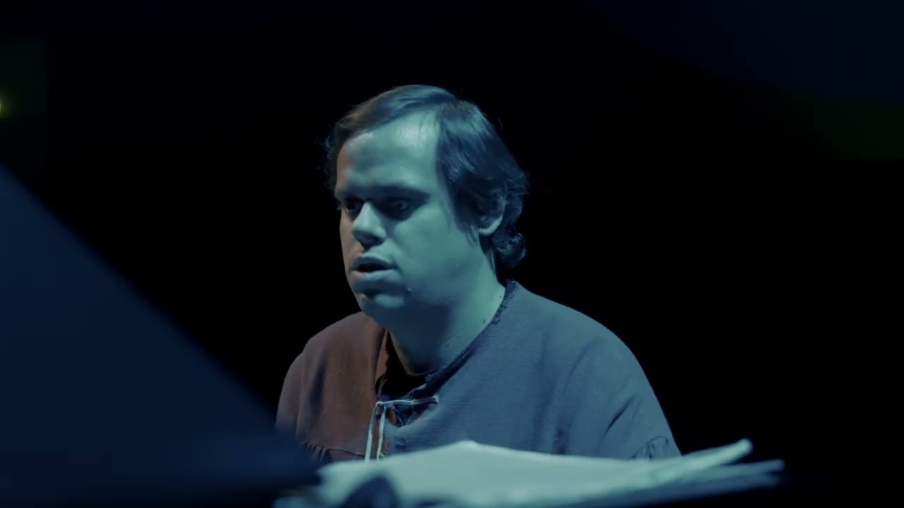 La imagen muestra a Ignasi, un hombre joven-adulto, sentado ante un piano con el tronco ligeramente inclinado hacia delante, en un escenario oscuro iluminado por una luz fría azulada que resalta su rostro y la parte superior del cuerpo. Presenta hombros relajados, cabeza levemente ladeada y rasgos faciales serenos, con la boca ligeramente entreabierta, signo de escucha atenta y concentración profunda. Su actitud es recogida e introspectiva, totalmente absorto en la interpretación musical. 