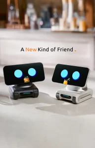 En esta imagen aparecen dos unidades de LOOI Robot situadas sobre una superficie clara y brillante, posiblemente una encimera de cocina o una mesa de mármol. El fondo está desenfocado con tonos cálidos, lo que genera un ambiente doméstico acogedor. El robot de la izquierda es negro y el de la derecha es blanco, mostrando las dos variantes de color. Ambos sostienen un smartphone en horizontal mostrando una cara animada con dos ojos redondos azul turquesa sobre fondo negro. En el robot negro, debajo de los ojos aparece una pequeña hamburguesa animada, mientras que en el robot blanco aparece un vaso con bebida y pajita. Sobre la escena, centrado en la parte superior, se lee el texto: “A New Kind of Friend.” con la palabra “New” destacada en color naranja.