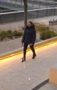 Imagen de una persona caminando por una pasarela urbana con bastón blanco de movilidad y Vector, rodeada de barandillas metálicas, jardineras y pavimento claro