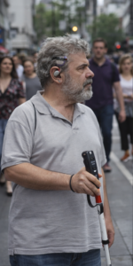 Un hombre de piel clara en una calle urbana, fotografiado de perfil, mirando hacia la izquierda.

Tiene el pelo canoso rizado y barba gris. Lleva una camiseta tipo polo gris y vaqueros azules. En la oreja izquierda lleva un dispositivo auditivo externo.

Sostiene un bastón blanco con una franja roja; la empuñadura es negra y tiene botones y una pequeña pantalla. Detrás se ven varias personas caminando, desenfocadas, y al fondo una calle con edificios.