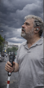 Un hombre de piel clara en una calle, en primer plano, mirando hacia arriba con expresión atenta.

Tiene el pelo canoso rizado y barba gris. Lleva un polo gris. En la oreja izquierda lleva un dispositivo auditivo externo con una pieza que rodea la oreja.

Sujeta un bastón blanco con una franja roja; en la parte superior hay una empuñadura negra con botones y una pequeña pantalla. En la pantalla se alcanza a leer “6.3 MP” y debajo “0.0 m”. El fondo muestra una calle con coches y edificios desenfocados, y un cielo muy oscuro y nublado.